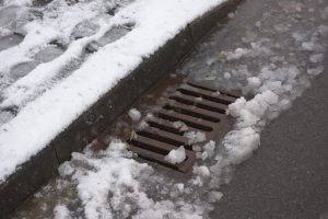 Side road drain partially covered with melted snow, slush, and ice after a winter storm.