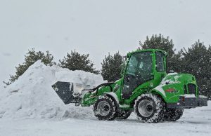 Snow plow truck pushing and piling snow in a commercial parking lot during snowfall.